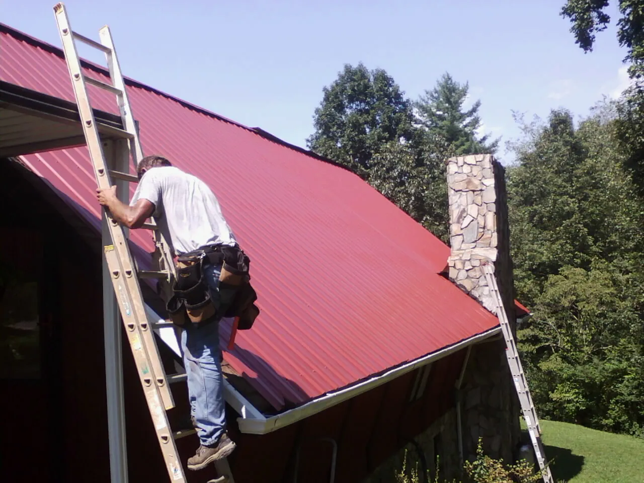 Roofer on a ladder beside a red metal roof and stone chimney — Soffit & Fascia Repair by Roofing Brothers in Normandy